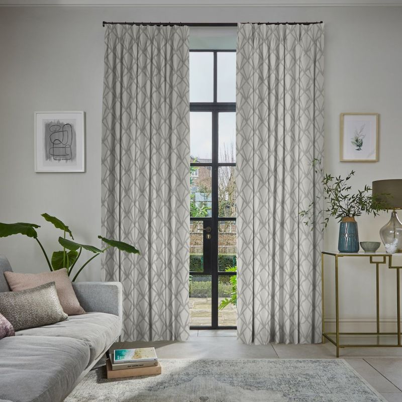 Patterned drapery panels, light grey with a geometric motif, hung on a rod and parted; filtering soft daylight through tall black-framed French doors into a living room overlooking a garden.