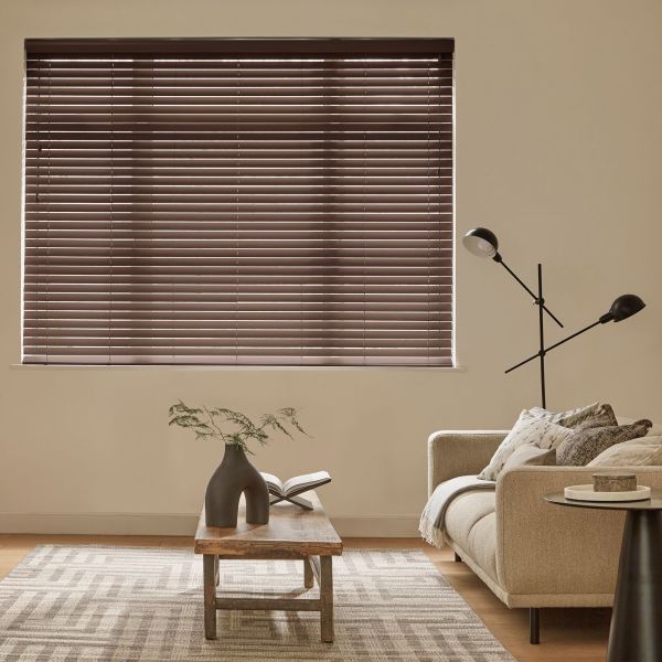Brown horizontal Venetian blinds covering a large window, fully closed and filtering soft daylight through narrow slats; a neutral living room with beige sofa, floor lamp, wooden coffee table and rug.