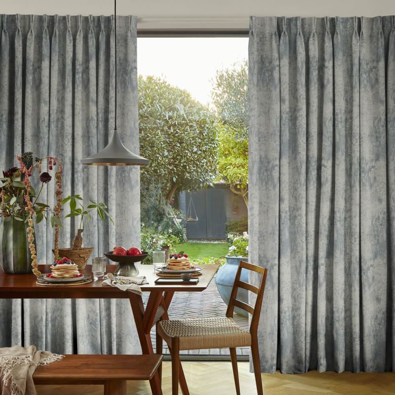 Pleated grey textured curtains, drawn aside to reveal a door, filtering soft daylight; a dining table set with pancakes and fruit, pendant lamp and chair, overlooking a sunlit garden.