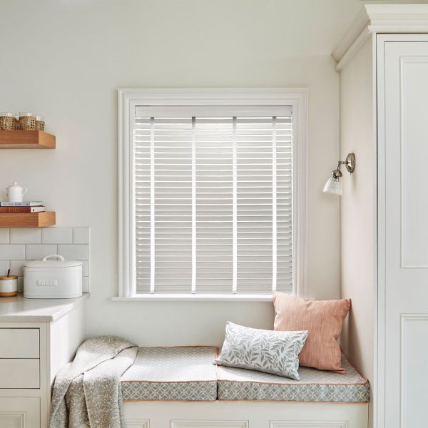 White horizontal Venetian blinds inside the window frame with narrow vertical tapes — closed, filtering soft daylight — above a cozy kitchen window seat with cushions, pale cabinetry, and a wall sconce.
BREAD