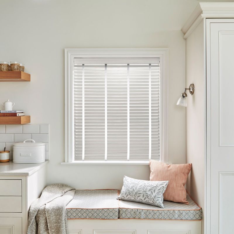 White horizontal Venetian blinds inside the window frame with narrow vertical tapes — closed, filtering soft daylight — above a cozy kitchen window seat with cushions, pale cabinetry, and a wall sconce.
BREAD