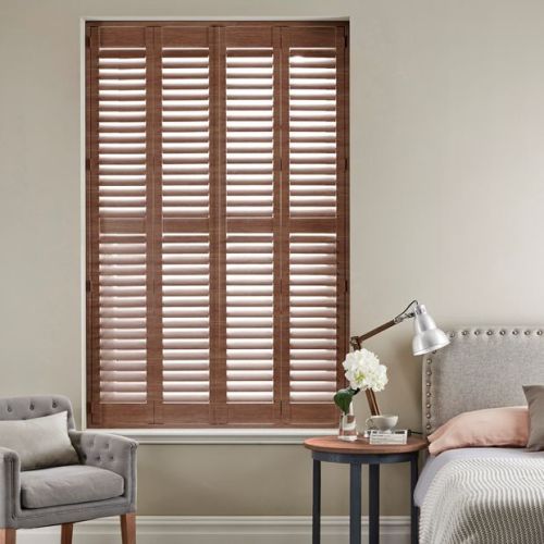 Wooden plantation shutters covering the full window, horizontal slats tilted to admit soft light; in a neutral-toned bedroom beside an upholstered bed, nightstand with lamp and chair.