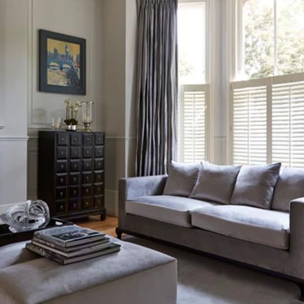 Floor-to-ceiling pleated gray curtains alongside white café-style louvered shutters covering a bay window, partially closed and filtering bright daylight; modern living room with gray sofa, ottoman, dark cabinet.