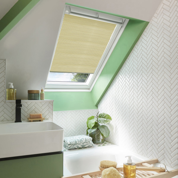 A pleated yellow skylight blind, mounted inside the frame, is partially lowered, filtering daylight over a sloped bathroom with green-painted trim, white herringbone tiles, sink, tub, and plant.
