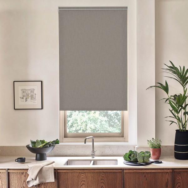 Roller blind covers most of a kitchen window above a stainless sink, with countertop, faucet, leafy plants, framed sketch on wall and wooden cabinets below.