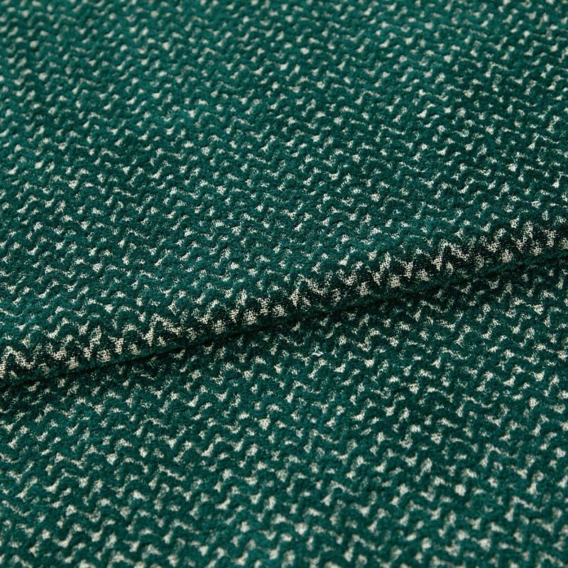 Green boucle-like fabric with dense cream zigzag pattern, folded into overlapping layers; close-up macro view on a neutral surface with soft, even lighting emphasizing the texture.