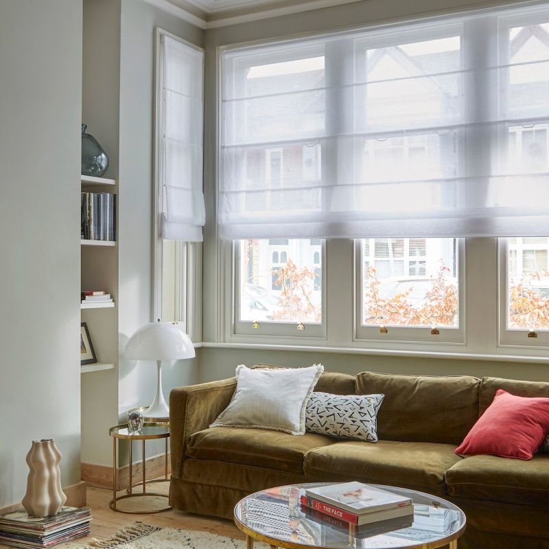 Sheer white Roman blinds lowered across a bay of windows, softly diffusing daylight; a living room with an olive velvet sofa, patterned and red cushions, round glass coffee table, books, and a white lamp.

THE FACE