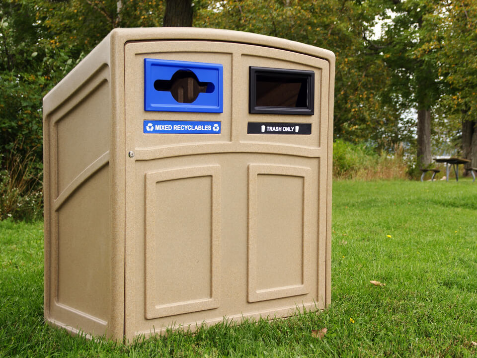 durable beige plastic roto-molded waste and recycling container with a sloped top and hinged front door located in a municipal park area