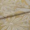 Botanical-patterned upholstery fabric, folded to show a layered edge; cream leaf-and-flower woven motif on mustard-yellow background, close-up studio shot with soft even lighting.