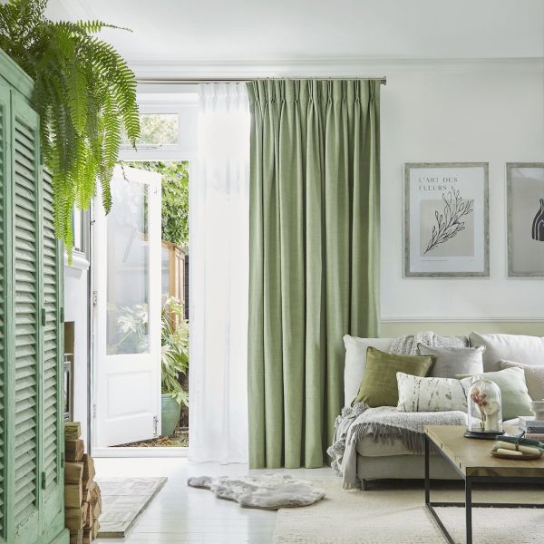 Green pinch-pleated curtains with an inner white sheer, partly drawn and filtering daylight beside an open white door. Bright, plant-filled living room with pale sofa, cushions, wooden coffee table, botanical wall art.
L'ART DES FLEURS