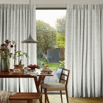 Floor-length, pleated pale curtains with subtle botanical print, parted at center; partially open, filtering daylight; framing dining table with pancakes, pendant lamp and wooden chairs, and a sunlit garden beyond.