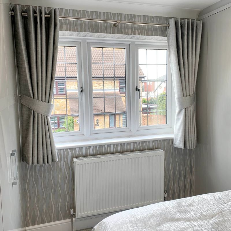 Grey pleated, eyelet-top curtains on a metal rod, pulled back with matching tiebacks, framing a threeâ€‘pane casement window and filtering daylight into a neutral bedroom with patterned wallpaper and white radiator.