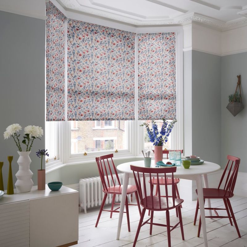 Patterned floral roller blinds covering a three-panel bay window, lowered mid-height and filtering daylight; in a pale-green dining room with a white round table, red chairs, and vases of flowers.
