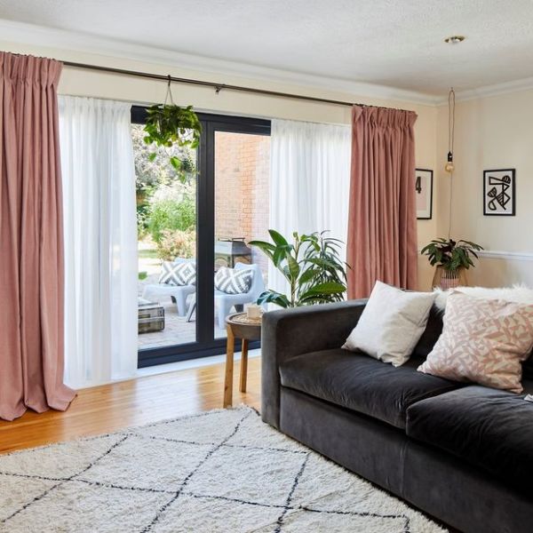 Floor-length blush-pink pleated curtains with sheer panels on a black rod, pulled to sides while sheers filter daylight; framing sliding glass doors to a sunny patio in a living room.