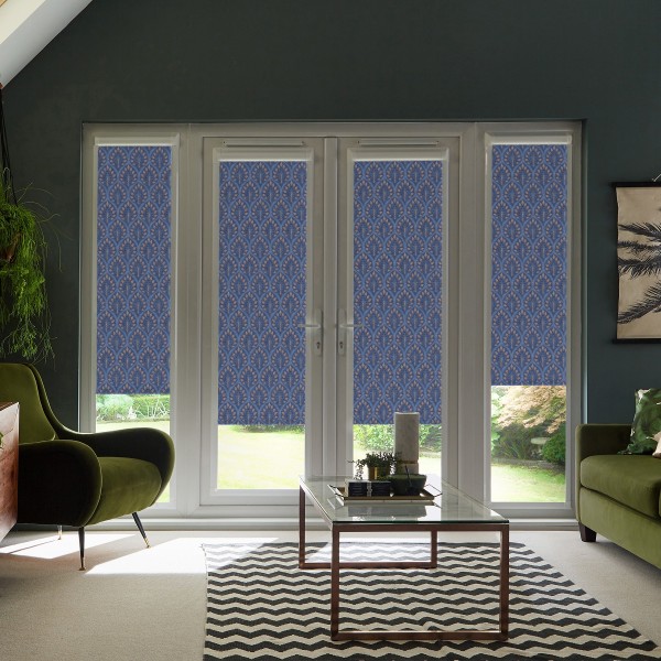 Blue patterned roller blinds cover lower halves of four tall glass doors; inside, a modern living room with velvet green chairs, glass coffee table, chevron rug and dark walls.