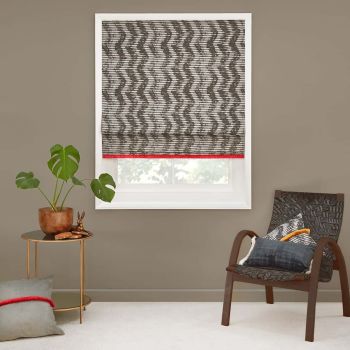 Roman-style patterned blind lowered over a white-framed window, black zigzag stripes with red trim, filtering daylight. Context: neutral-toned living-room corner with potted plant, round side table and woven chair.
