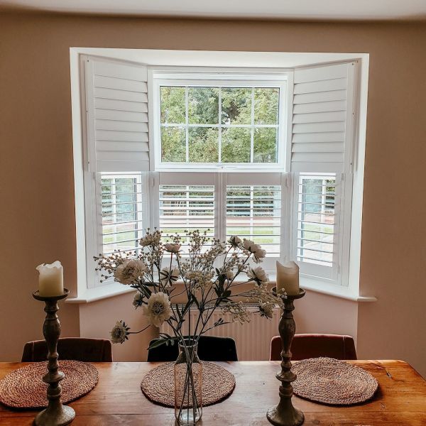 White plantation shutters on a bay window — top panels folded outward, lower louvers partially closed — filtering soft daylight, framing a leafy garden over a dining table with candles and flowers.