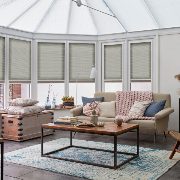 Light-grey honeycomb blinds lowered over tall conservatory windows, filtering soft daylight; living room with beige sofa draped in a pink chunky-knit throw, wooden coffee table and blue rug.