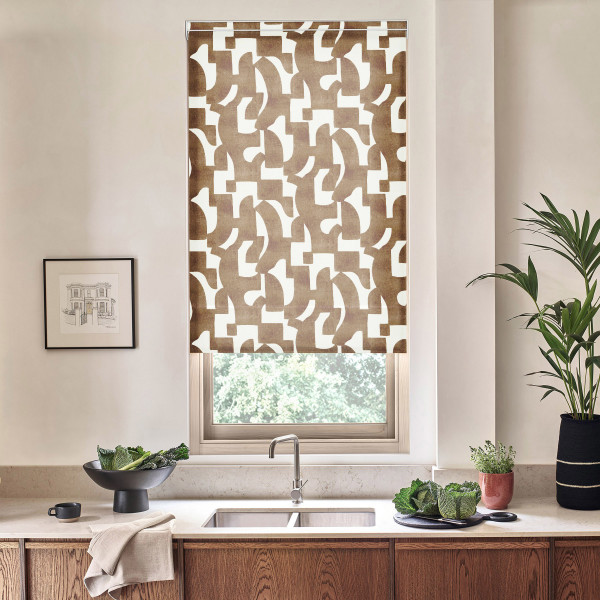 Patterned roller blind hangs over a window, filtering light above stainless sink; surrounding modern kitchen countertop features wooden cabinets, black bowl with greens, potted plants and framed sketch.