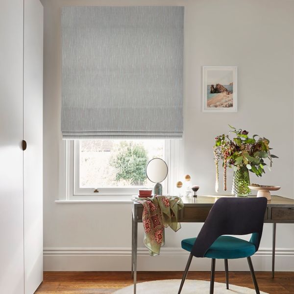 Light-blue patterned Roman blind lowered over a sash window, partially filtering daylight; sits above a wooden desk with a teal chair, mirror, vase of flowers and lit home-office interior.