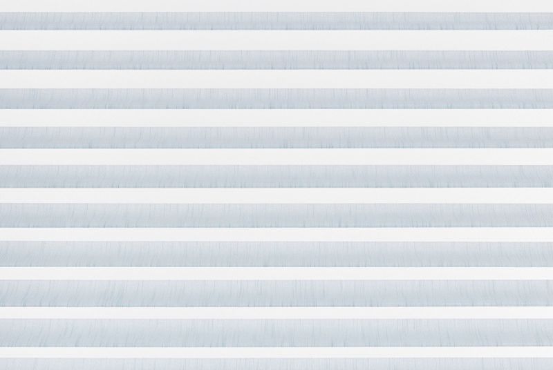 Horizontal zebra blinds with alternating white opaque and pale blue translucent fabric filling the frame, closed and filtering soft daylight, in a minimal interior with no visible view.
