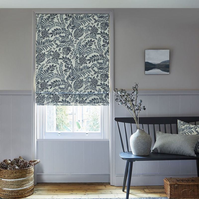 Patterned Roman blind lowered over a sash window, blue botanical print, partially covering glass and softly filtering daylight; hallway alcove with painted paneling, bench, ceramic vase, wicker basket, muted decor.