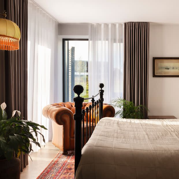 Sheer white curtains with flanking heavy greyâ€‘brown drapes, floorâ€‘length and partially drawn, filtering soft daylight through a glass door; bedroom contains an iron bed, tufted leather armchair, plants, rug.