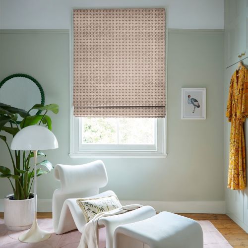 Patterned beige Roman blind lowered over central window, partially closed and softening daylight through a slim gap; in pastel-green reading nook with sculptural chair, plant, lamp, bird print and yellow robe.