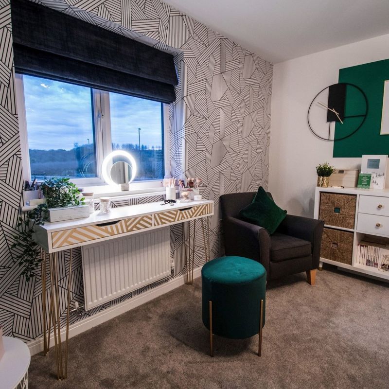 Dark charcoal Roman blind, partially lowered over a twin-pane window, gently filtering dusk light; occupies a patterned wallpapered bedroom/vanity corner with illuminated ring lamp, armchair, teal ottoman, soft carpet.