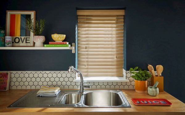 Beige wooden Venetian blinds centered in frame, closed and filtering warm daylight through slats; above a stainless double sink in a dark-blue kitchen with hex-tile backsplash, shelf, plants, utensils.
LOVE
JUICE