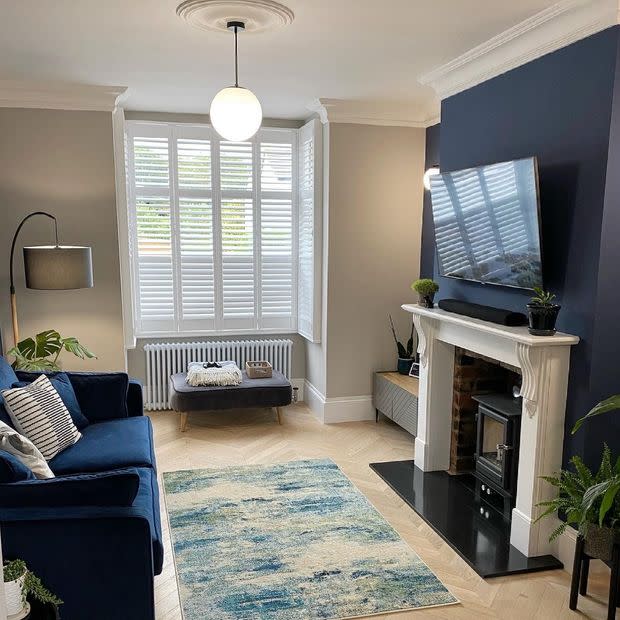White plantation shutters on a bay window, closed with angled louvers filtering soft daylight; living room features navy accent wall, fireplace, blue sofa, patterned rug, hardwood floor, potted plants.