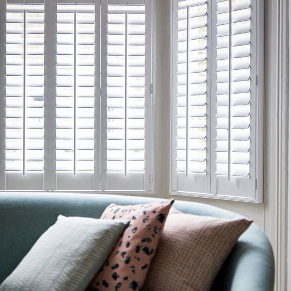 White plantation shutters covering corner bay windows, louvered panels tilted to diffuse bright daylight; in front, a curved teal sofa with patterned cushions in a softly lit living room.