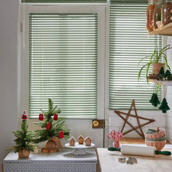 Horizontal pale-green Venetian blinds on a door and adjacent window, closed and softly filtering daylight; kitchen scene with small tabletop Christmas trees, gingerbread houses, candy canes and baking tools.