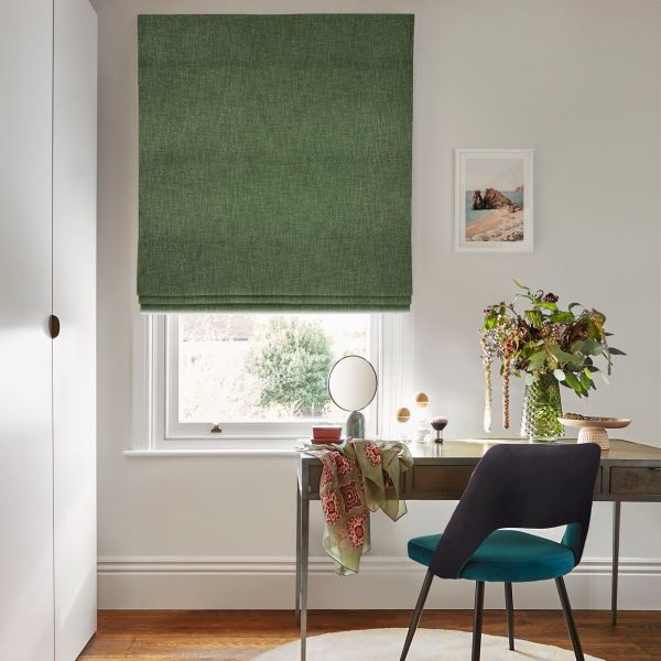 Green Roman blind, lowered with subtle folds, filtering soft daylight through a narrow bottom gap; above a desk with mirror, chair and vase of flowers in a bright home office.