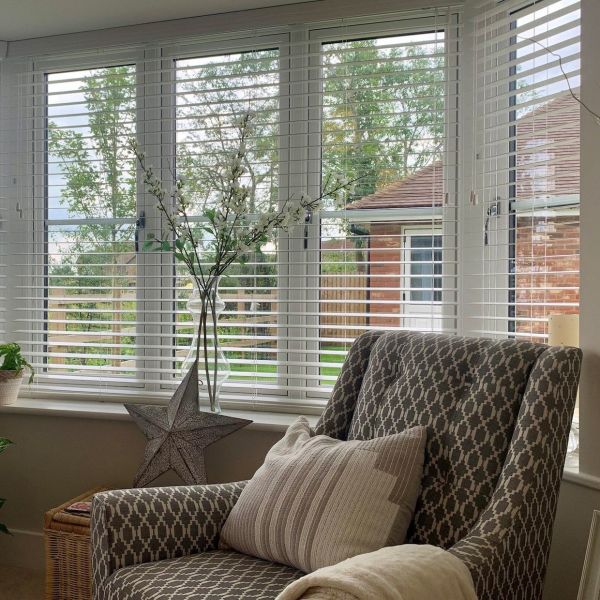 White horizontal Venetian blinds over a bay window, slats tilted partially open filtering daylight and framing a garden view; cozy living-room corner with patterned armchair, glass vase and metal star ornament.