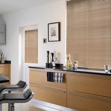Beige horizontal Venetian blinds covering two windows, fully lowered and closed, filtering soft daylight; foreground oak kitchen cabinets and black countertop with utensils, glass jars, and two black bar stools.
