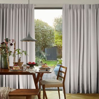 Pleated light-gray curtains, full-length and hung high, parted centrally—partially drawn and filtering daylight—framing a breakfast dining table with pancakes and a sunlit garden beyond with trees and a shed.