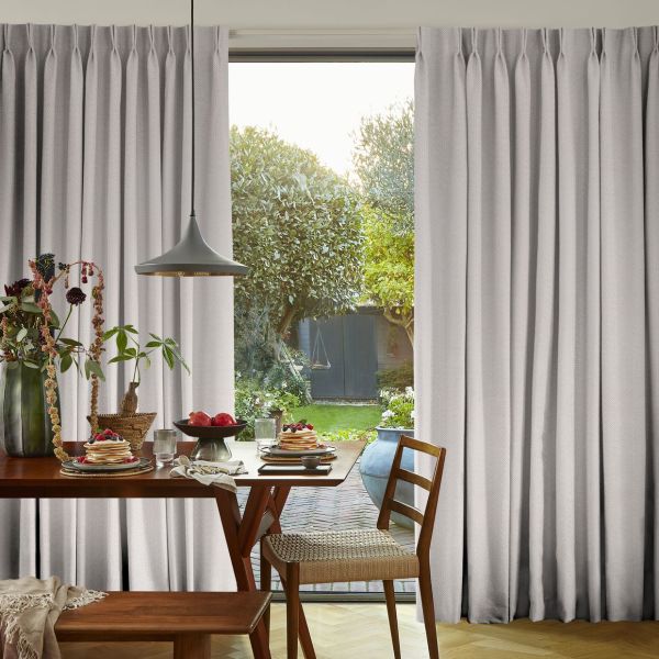 Pleated light-gray curtains, full-length and hung high, parted centrally—partially drawn and filtering daylight—framing a breakfast dining table with pancakes and a sunlit garden beyond with trees and a shed.