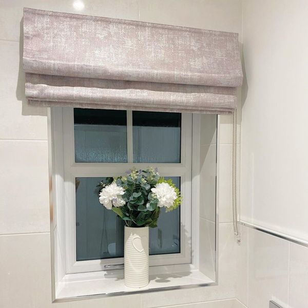 Light-pink Roman blind, folded with a bead-chain at the side; partially lowered over the top pane. Tiled bathroom, frosted window and ceramic vase with white flowers on the sill.