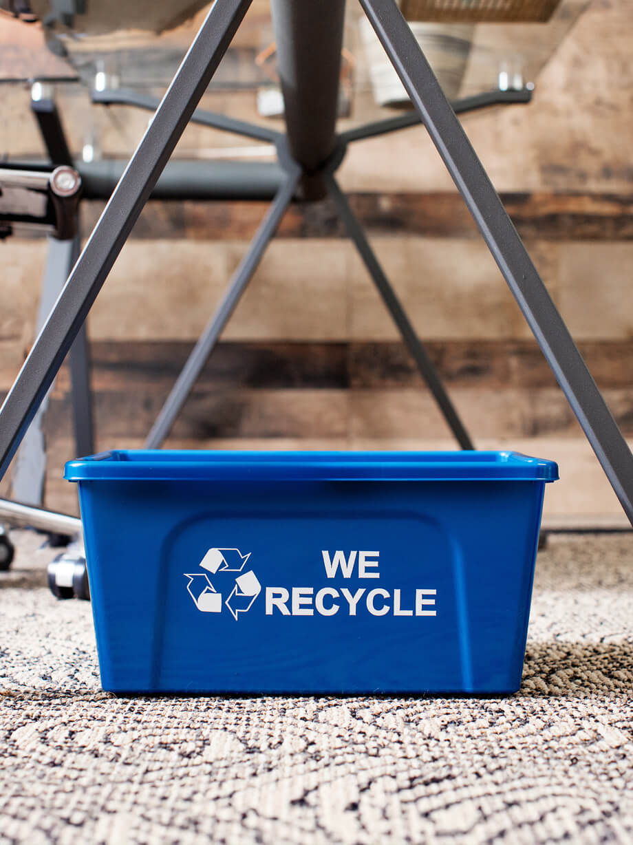blue deskside recycling container with we recycle stamped on the bin sitting under a modern office desk