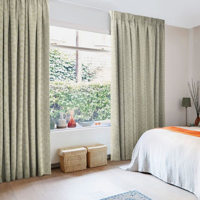 Pale green patterned pleated curtains, floor-to-ceiling and partially drawn, framing a wide sash window and filtering soft daylight; bedroom interior with bed, wicker baskets and leafy garden outside.