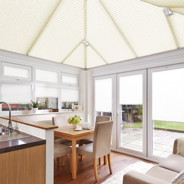 Cream pleated (cellular) blinds covering the pitched conservatory roof and windows, closed and diffusing bright daylight; small dining area with wooden table, upholstered chairs, kitchen sink and patio view.