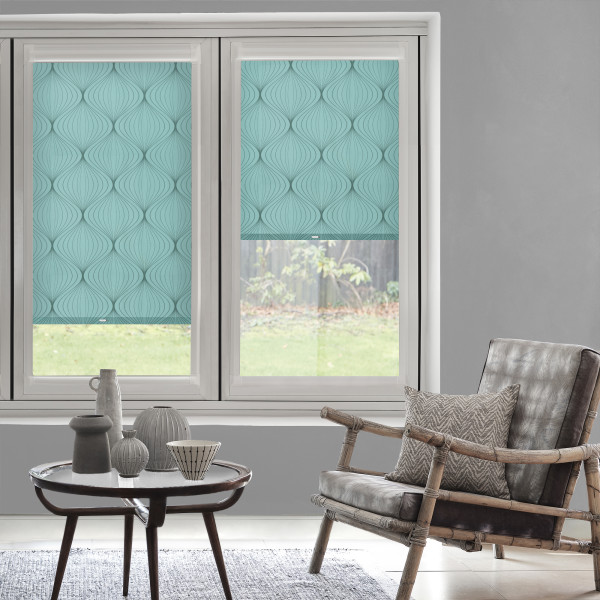 Turquoise, leaf-patterned roller blinds covering two sash windows—one fully lowered, one halfway—filtering soft daylight; living-room setting with gray walls, rattan armchair, patterned cushion, wooden coffee table and ceramics, garden view.