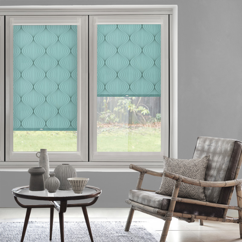 Turquoise, leaf-patterned roller blinds covering two sash windowsâ€”one fully lowered, one halfwayâ€”filtering soft daylight; living-room setting with gray walls, rattan armchair, patterned cushion, wooden coffee table and ceramics, garden view.