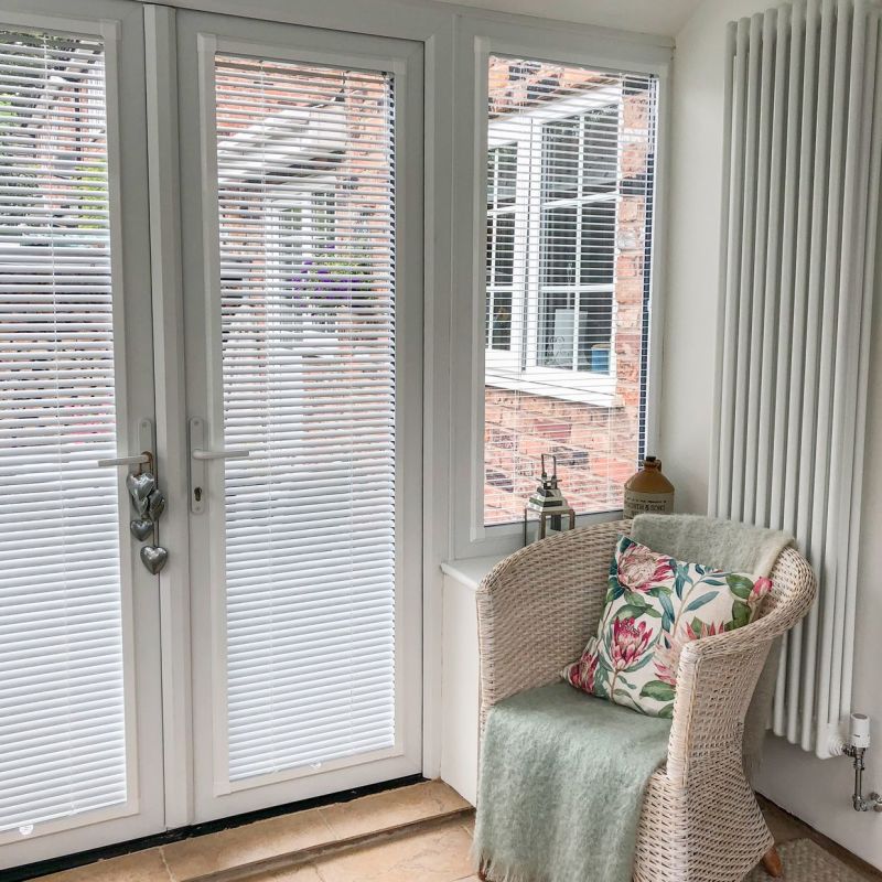 White horizontal Venetian blinds on double French doors and side window, closed with slats tilted to filter light; sunroom corner with wicker chair, floral cushion, radiator, tiled floor, brick exterior.