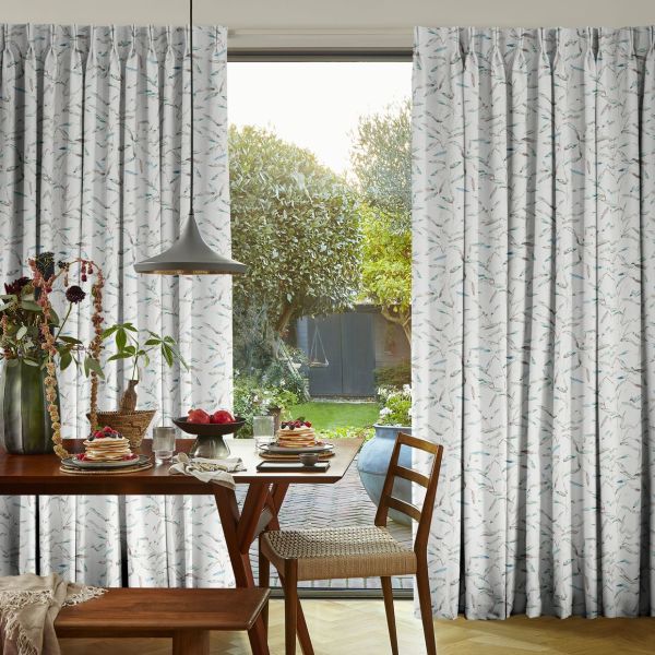 Floor-length pleated curtains patterned with pale leaves, pulled aside to frame a central opening over sliding doors; revealing a sunlit garden beyond and a breakfast-laid wooden dining table.