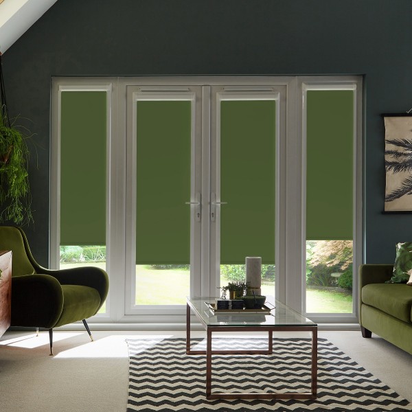 Four glass patio doors with olive-green roller blinds are closed, blocking most view; set in a dark-walled living room with green armchairs, glass coffee table, chevron rug, and plants.