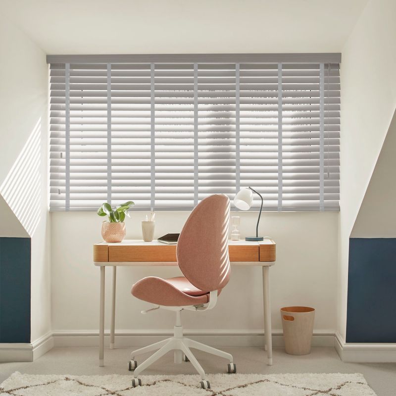 Horizontal venetian blinds drawn across a wide dormer window, slats partially tilted to filter sunlight; small home office alcove with wooden desk, pink swivel chair, lamp and plant.
