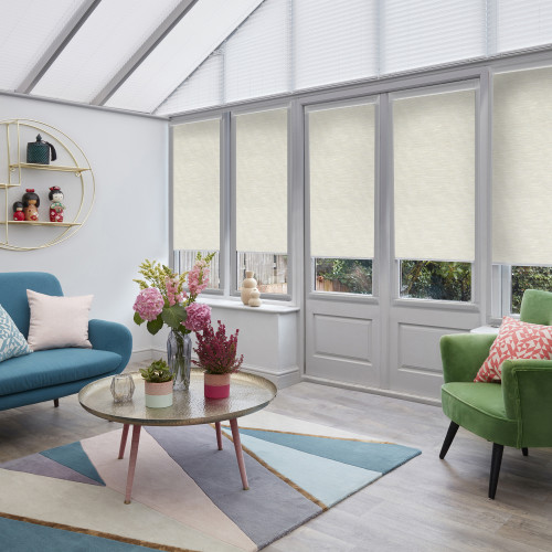 Beige roller blinds and pleated roof blinds: lowered across tall conservatory windows and doors, filtering daylight; sunroom with teal sofa, green armchair, round coffee table and floral centerpiece, garden view.