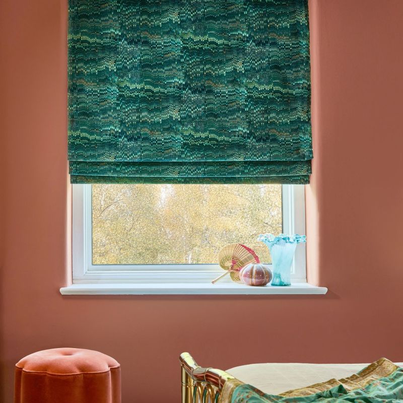Green, textured Roman blind, lowered halfway with folded tiers, filtering soft daylight through the window. Context: terracotta-painted bedroom walls, white sill holding a blue glass vase and small decorative items.
